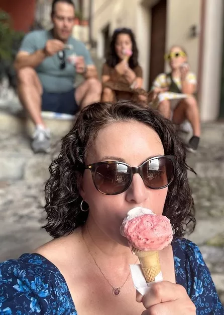 Woman from Adventurous Tastes sitting on steps eating gelato in Italy with her husband and two daughters blurry behind her.