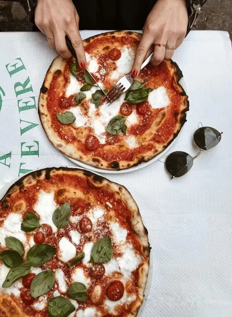 All visits to Rome with kids should include pizza! Overhead view of two pizzas on a table with hands cutting a slice.