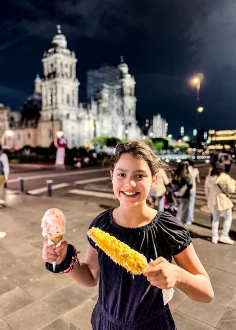 Street food is an essential part of visiting Mexico City with kids. A little girl holds corn on the cob and an ice cream cone at night near a Mexico City square