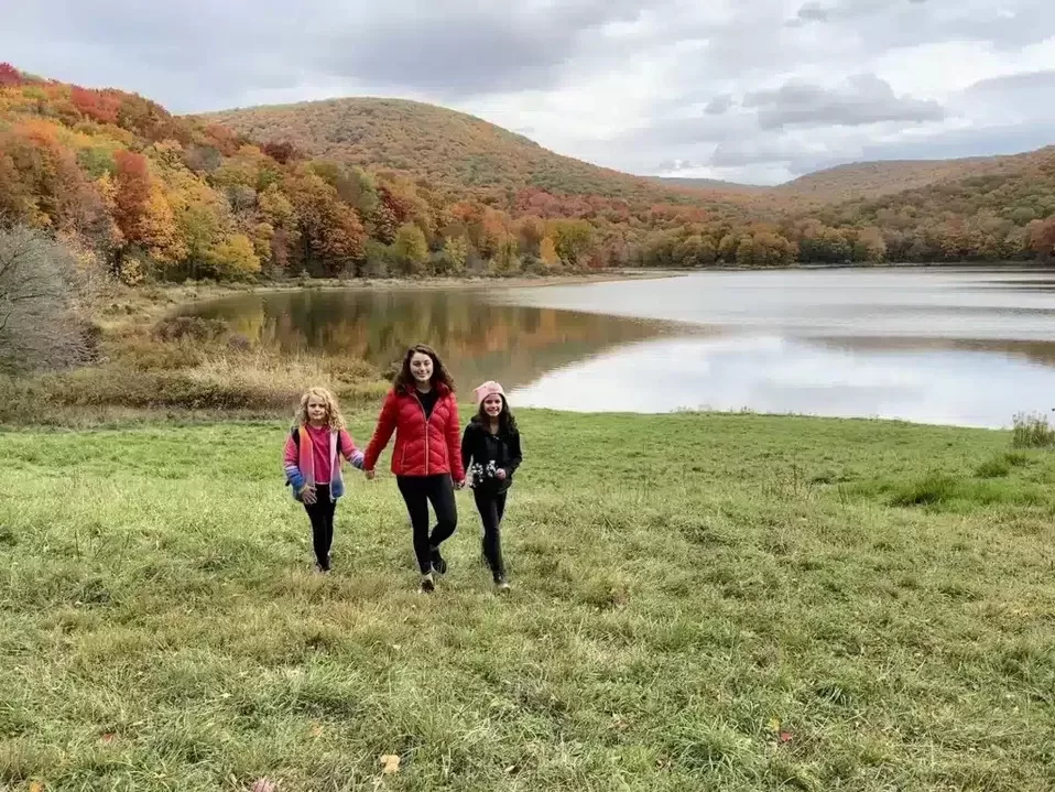 Fall break trip to the Catskills - Mother and two daughters from Adventurous Tastes holding hands near Alder Lake in the fall with trees and lake behind them